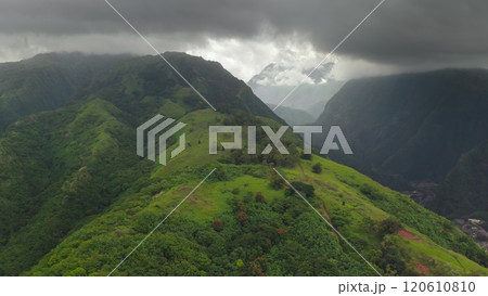 Aerial view captures breathtaking beauty of Tahiti's landscape, lush green valleys hills, dramatic mountain peaks, cloudy moody sky, small village houses in canyon. Remote travel to French Polynesia 120610810