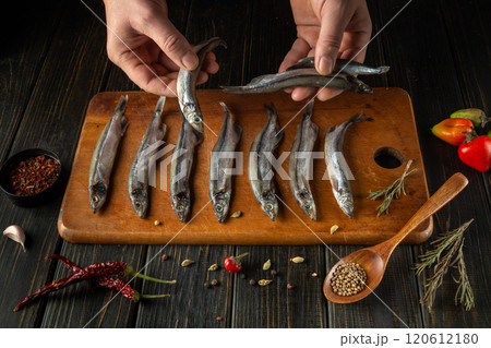 Hands carefully arrange fresh fish on a wooden cutting board with herbs, spices, and colorful peppers nearby, showcasing the preparation of a delightful culinary dish 120612180