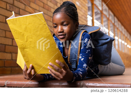 In school, african american girl lying on floor reading yellow notebook with backpack on 120614288