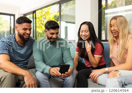 diverse friends sitting on couch, laughing and looking at smartphone together 120614564