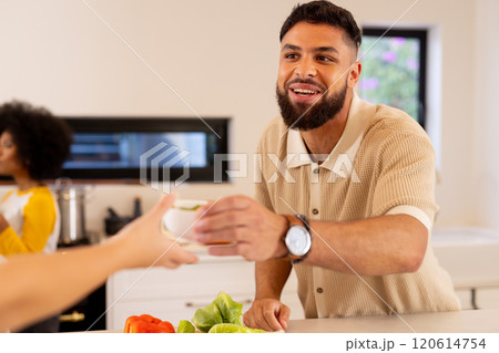 Smiling man in kitchen handing cup to friend while preparing vegetables 120614754