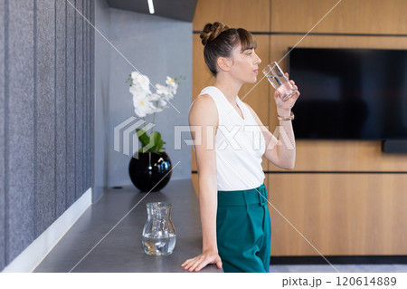 Drinking water from glass, woman standing in modern office with pitcher nearby 120614889