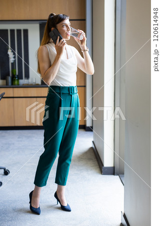 Talking on smartphone and drinking water, businesswoman standing by office window Talking on smartphone and drinking water, businesswoman standing by office window 120614948