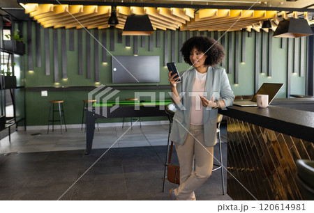 Using smartphone and smiling, businesswoman in modern office with laptop and coffee, copy space 120614981