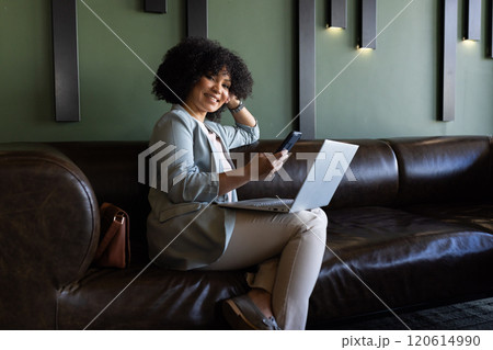 Using smartphone and laptop, businesswoman working remotely on leather couch 120614990