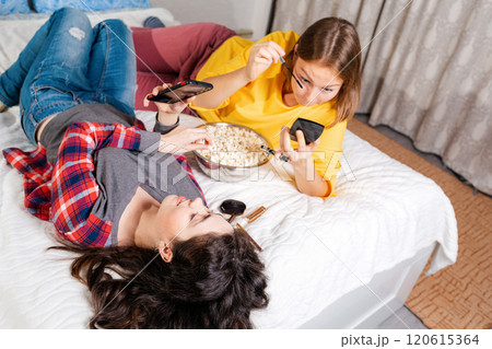 Two young women are lying on the bed, eating popcorn, using smarfton, and decorative cosmetics are lying next to them. Top view.Concept of LGBT relationships and recreation at home Two young women are lying on the bed, eating popcorn, using smarfton, and decorative cosmetics are lying next to them. Top view.Concept of LGBT relationships and recreation at home 120615364