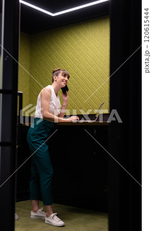 Talking on smartphone, woman working on laptop in modern office space 120615444