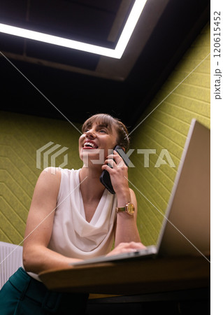 Talking on smartphone, businesswoman smiling and using laptop in office 120615452