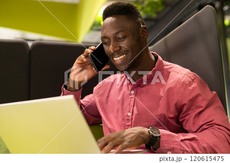Smiling african american man using laptop and talking on smartphone in modern office 120615475