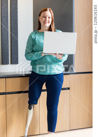 Holding laptop, woman with prosthetic leg smiling in modern office 120616165