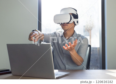 At office, Using VR headset, asian man interacting with virtual environment while working on laptop 120616182