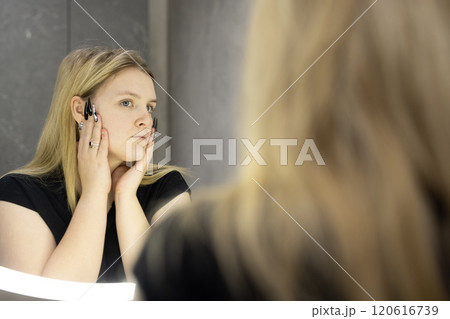 A young blonde woman examines herself in a modern bathroom mirror with illuminated edges, adjusting her hair with a focused expression. A young blonde woman examines herself in a modern bathroom mirror with illuminated edges, adjusting her hair with a focused expression. 120616739