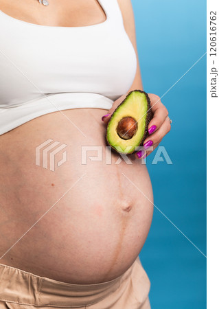 Close-up of a pregnant belly with an avocado held by hand against a blue background Close-up of a pregnant belly with an avocado held by hand against a blue background 120616762