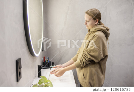 A young woman in a hoodie rinses her hands under running water at a modern sink in a minimalistic bathroom with a circular illuminated mirror. 120616783