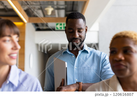 Holding laptop, african american man checking time on watch while standing with colleagues 120616807