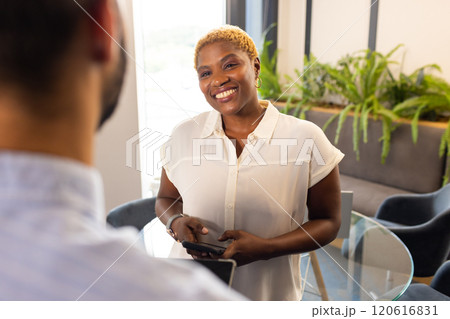 Smiling woman holding smartphone, engaging in conversation with colleague in office 120616831