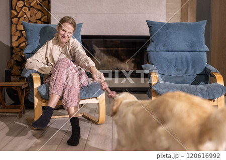 A cheerful young woman plays tug-of-war with her Golden Retriever in a cozy living room, sitting in an armchair near a fireplace. A cheerful young woman plays tug-of-war with her Golden Retriever in a cozy living room, sitting in an armchair near a fireplace. 120616992