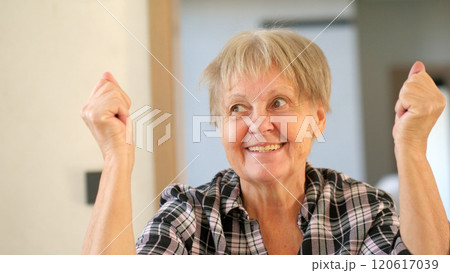 Joyful elderly woman with a triumphant expression, raising her fists in a celebratory gesture indoors. Joyful elderly woman with a triumphant expression, raising her fists in a celebratory gesture indoors. 120617039