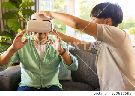 Using VR headset, asian father experiencing virtual reality with assistance in living room 120617593