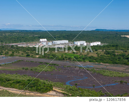 Aerial view captures industrial facility amid lush greenery, wetlands, and distant mountains under bright sky. 120617856