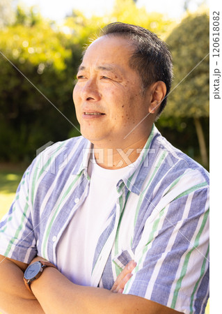 Smiling asian middle-aged man standing outdoors, crossing arms and looking away, enjoying sunny day 120618082