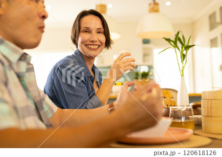 At home, Smiling middle-aged woman enjoying meal with family, holding glass at dining table 120618126