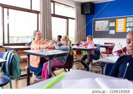 Female teacher assisting young girl with schoolwork in classroom, other diverse students studying Female teacher assisting young girl with schoolwork in classroom, other diverse students studying 120618368