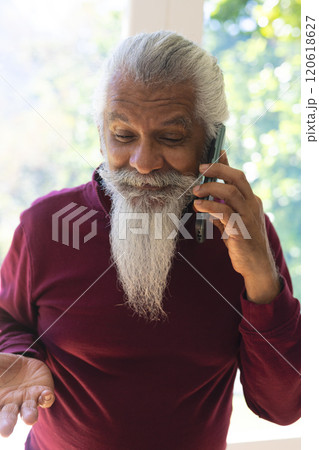 Happy biracial senior man with long beard, using smartphone in sunny living room, copy space 120618627