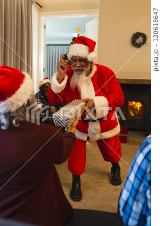 Happy biracial man in santa clothes giving gift to friend next to christmas tree at home Happy biracial man in santa clothes giving gift to friend next to christmas tree at home 120618647