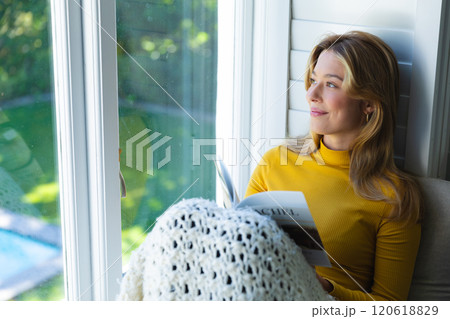 Happy caucasian woman sitting on couch with book and looking out window in sunny room at home 120618829