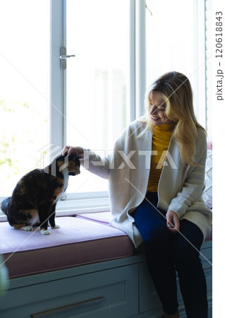 Happy caucasian woman sitting on windowsill and stroking cat in sunny room at home 120618843