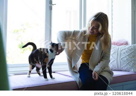 Happy caucasian woman sitting on windowsill and stroking cat in sunny room at home Happy caucasian woman sitting on windowsill and stroking cat in sunny room at home 120618844