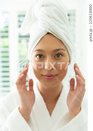 Happy caucasian woman in bathrobe looking in mirror, touching face and smiling in sunny bathroom Happy caucasian woman in bathrobe looking in mirror, touching face and smiling in sunny bathroom 120618860