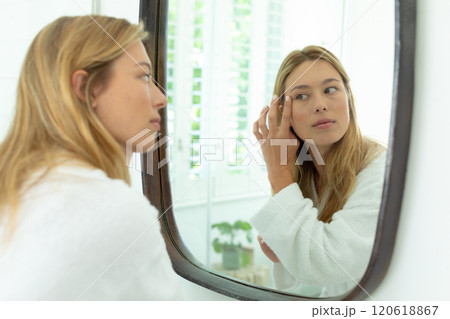 Happy caucasian woman in bathrobe looking in mirror and touching face in sunny bathroom 120618867