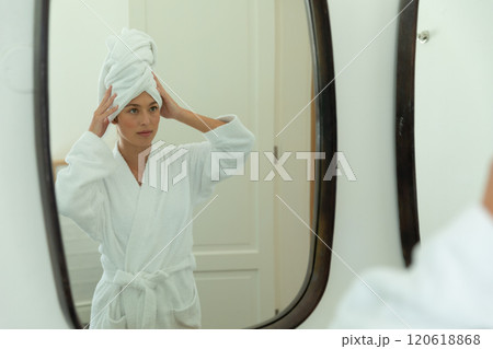 Happy caucasian woman in bathrobe looking in mirror and holding towel on head in sunny bathroom 120618868
