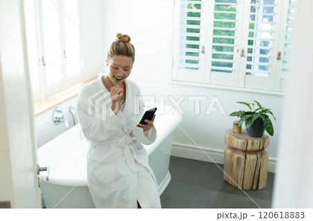 Happy caucasian woman in bathrobe brushing teeth and using smartphone in sunny bathroom 120618883