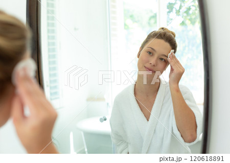 Happy caucasian woman in bathrobe cleaning face with cotton pad in sunny bathroom 120618891