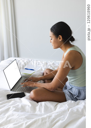 Busy biracial woman with smartphone and notebook using laptop with copy space in bedroom Busy biracial woman with smartphone and notebook using laptop with copy space in bedroom 120618960