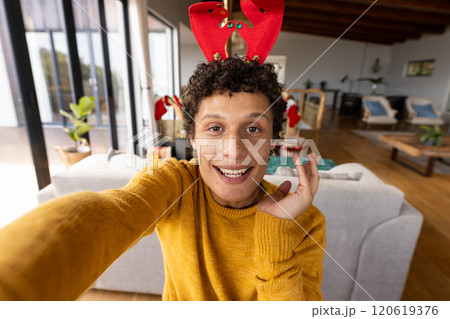 Portrait of happy biracial man wearing reindeer antlers and having video at christmas at home 120619376