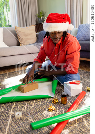 Happy african american man in christmas hat sitting on floor wrapping gifts, copy space 120619489