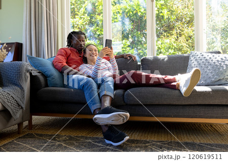 Happy diverse couple using smartphone together relaxing on couch in sunny living room 120619511