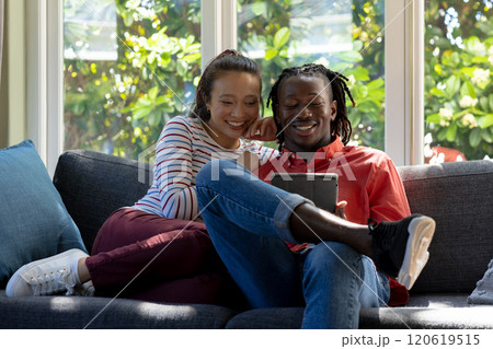 Happy diverse couple using tablet together sitting on couch smiling in sunny living room Happy diverse couple using tablet together sitting on couch smiling in sunny living room 120619515
