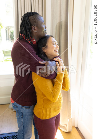 Happy diverse couple standing by window embracing and smiling in sunny living room, copy space 120619560