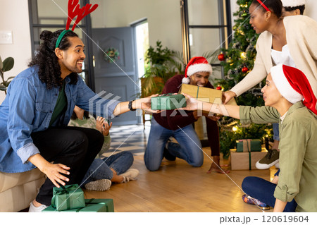 Wearing festive hats, multiracial friends exchanging gifts by christmas tree, at home 120619604