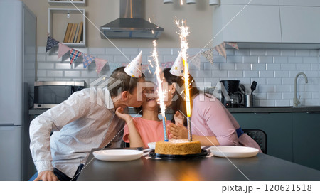Father and mother in festive hats kiss their daughter while admiring the sparkling ice fountains on a birthday cake Father and mother in festive hats kiss their daughter while admiring the sparkling ice fountains on a birthday cake 120621518