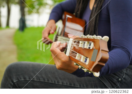 Woman with Brown Skin and Long Black Hair with Dreadlocks Sitting on Bench Playing Guitar in Green 120622362