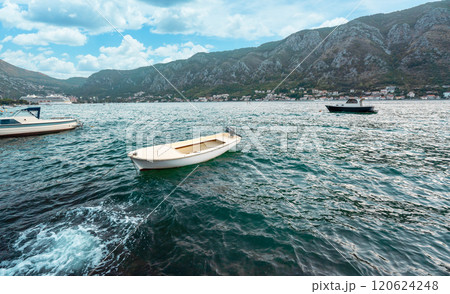 A small white boat floating on a serene bay surrounded by mountains, with rippling water under a vibrant blue sky. 120624248