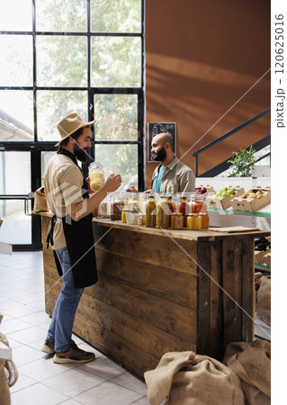 Caucasian storekeeper wearing a hat and black apron holds a jar filled with organic food item. Customer looks around on the other side of counter while vendor arranges glass containers. Caucasian storekeeper wearing a hat and black apron holds a jar filled with organic food item. Customer looks around on the other side of counter while vendor arranges glass containers. 120625016