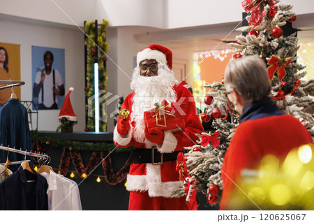 Joyful worker in costume carries raffle in clothing store, inviting senior woman to participate at contest and win free clothes during christmas season. Festive decorated shopping center. Joyful worker in costume carries raffle in clothing store, inviting senior woman to participate at contest and win free clothes during christmas season. Festive decorated shopping center. 120625067