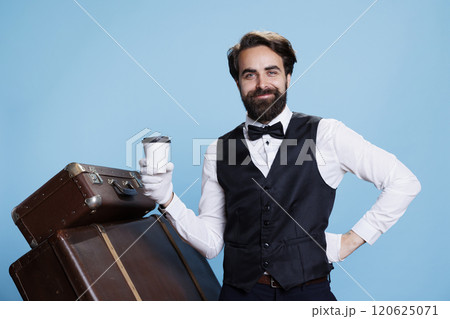 Positive doorman acting optimistic in studio, enjoying cup of coffee and standing next to luggage. Classy young hotel porter wearing formal attire and drinking tea as beverage. 120625071